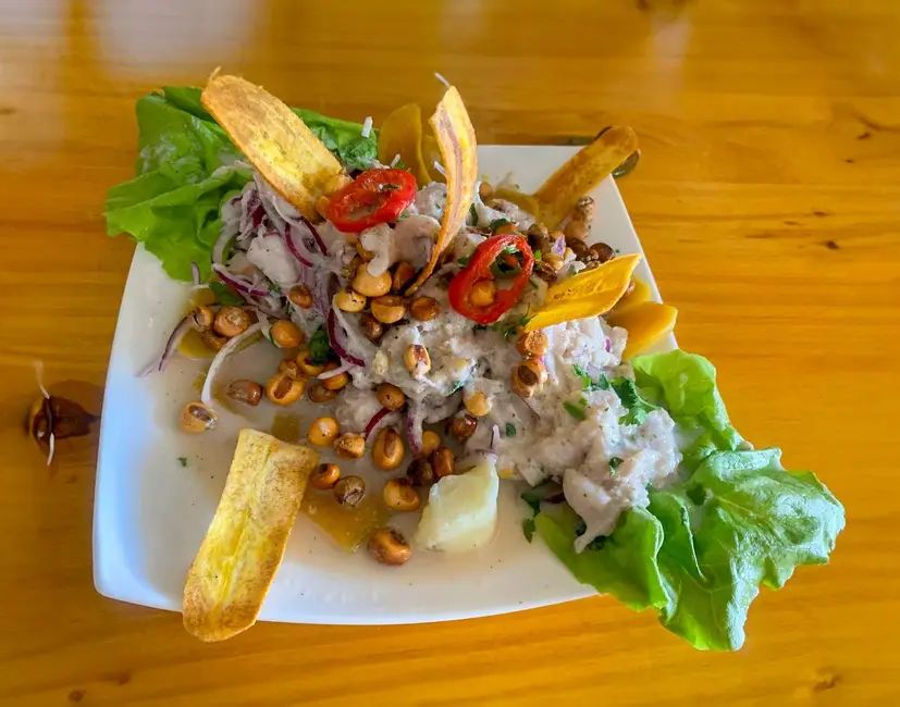 El ceviche de Maggy’s Chifa en plato blanco sobre una mesa de madera: pescado, cebolla morada, ají, cancha, lechuga y chifles de plátano.